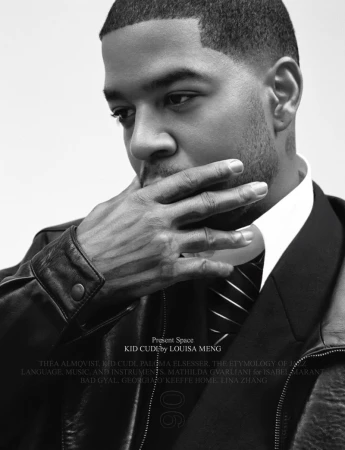 Black and white portrait of Kid Cudi in a leather jacket and tie, hand to face, gazing downward against a neutral background.