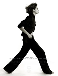 Mathilda Gvarliani walking in profile wearing a dark jacket and wide-leg trousers against a white seamless studio background.