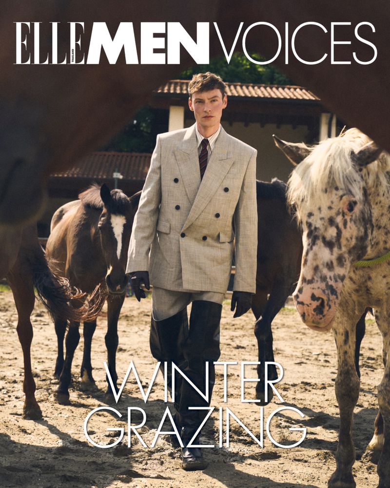 A man in a grey suit stands among horses in a sunny outdoor environment. The image is an editorial fashion shot with the Elle Men Voices logo.
