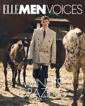 Man wearing a tailored double-breasted suit standing amongst several horses outdoors in an earthen paddock
