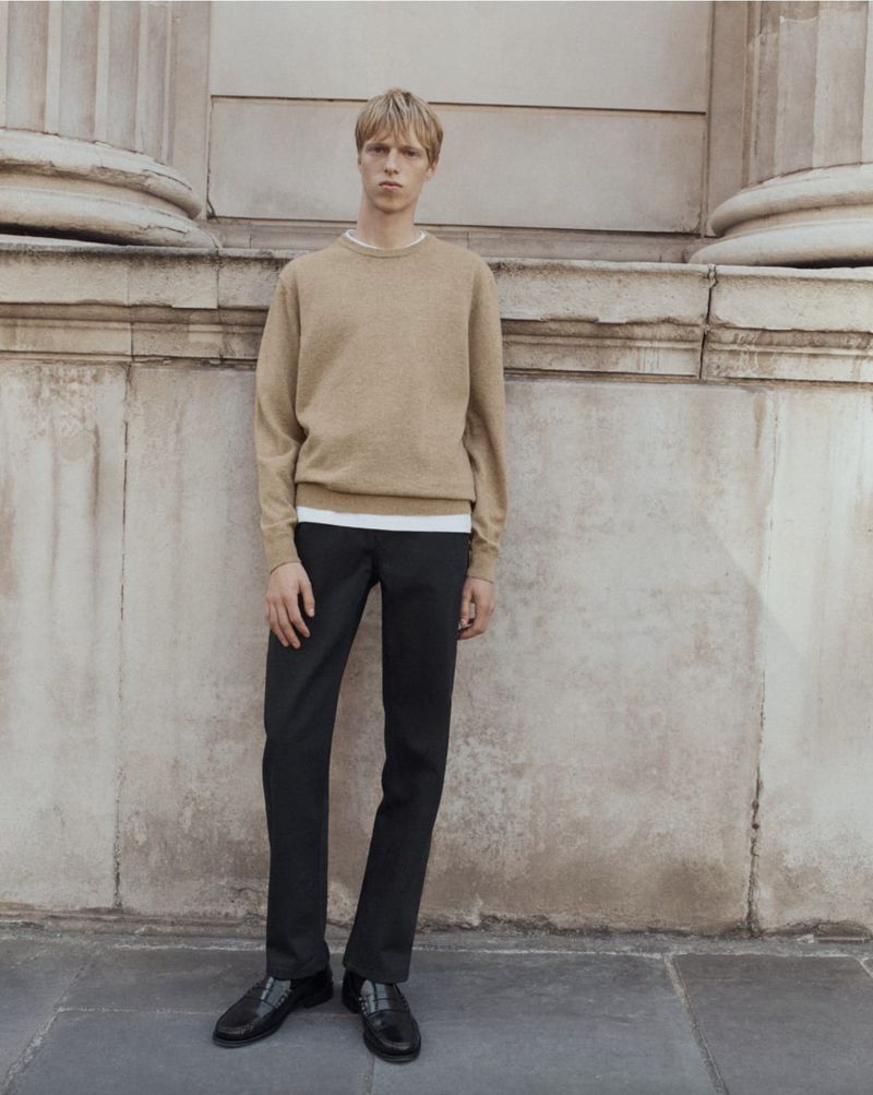 Man in a tan sweater and black pants stands against a textured wall with architectural columns. Full shot captures his casual pose.