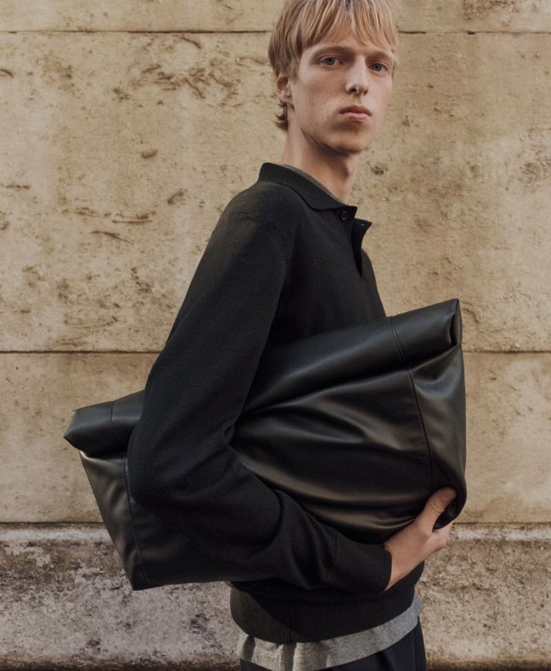 An adult male is holding a large black leather bag, posing in front of a textured wall in a fashion editorial style, medium studio shot.