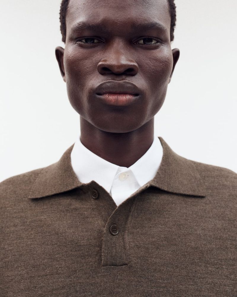 Portrait of a dark-skinned adult man in a brown knit polo shirt with a white collared shirt underneath, against a white backdrop.