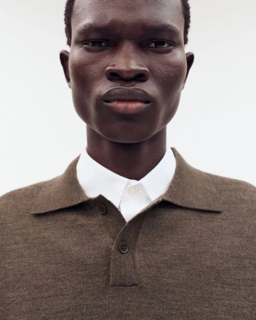 Portrait of a dark-skinned adult man in a brown knit polo shirt with a white collared shirt underneath, against a white backdrop.