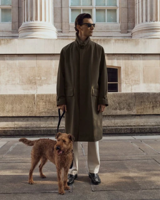 Stylish man in sunglasses and a long coat walking an Irish Terrier dog outdoors in front of a classical-style building with columns.