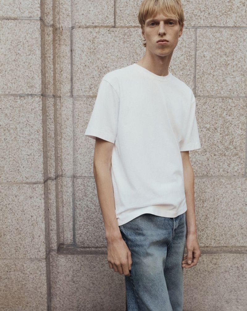 Model wearing a white t-shirt and blue jeans standing against a textured stone block wall