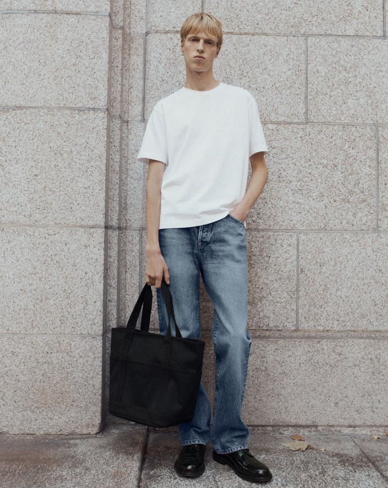 Man in white t-shirt and jeans holds black tote bag standing against a stone wall, with a neutral expression in an outdoor setting.