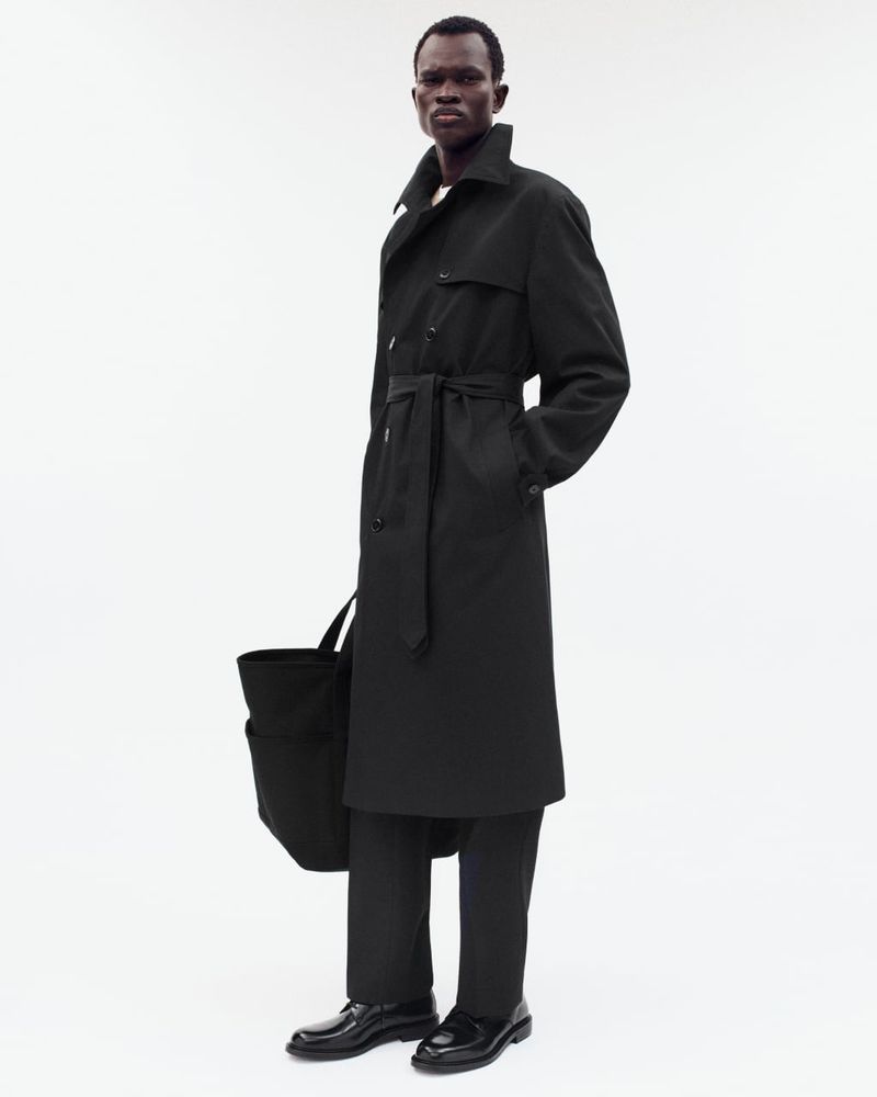 A full shot captures a black man in a black trench coat and pants, holding a tote bag against a bright, neutral backdrop.