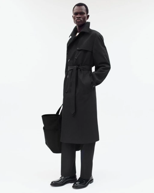 A full shot captures a black man in a black trench coat and pants, holding a tote bag against a bright, neutral backdrop.