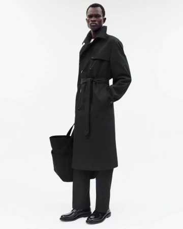 A full shot captures a black man in a black trench coat and pants, holding a tote bag against a bright, neutral backdrop.