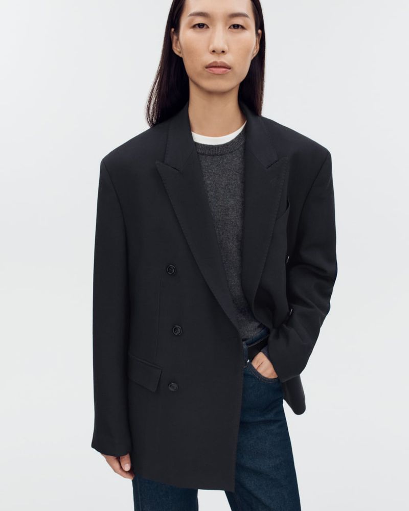 An Asian woman in her 20s poses in a black, oversized double-breasted blazer, dark jeans, and a gray sweater against a white background.