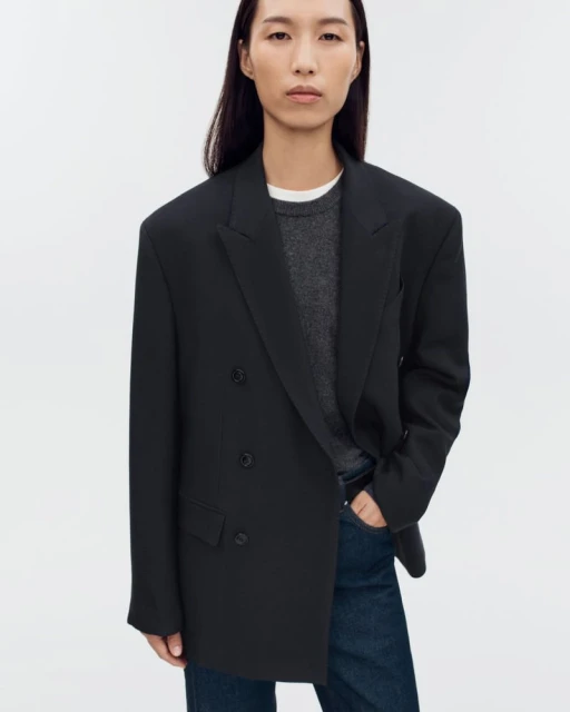An Asian woman in her 20s poses in a black, oversized double-breasted blazer, dark jeans, and a gray sweater against a white background.