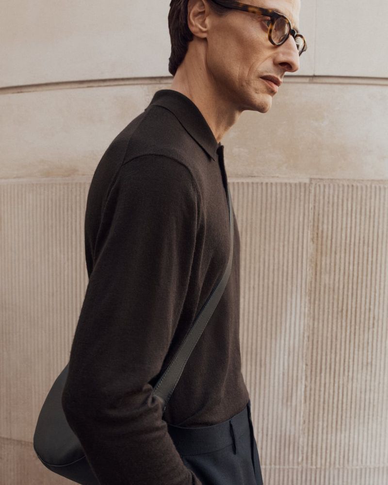 A stylish adult man with glasses in a brown polo shirt, wearing a dark shoulder bag walks past a light-colored ridged wall.