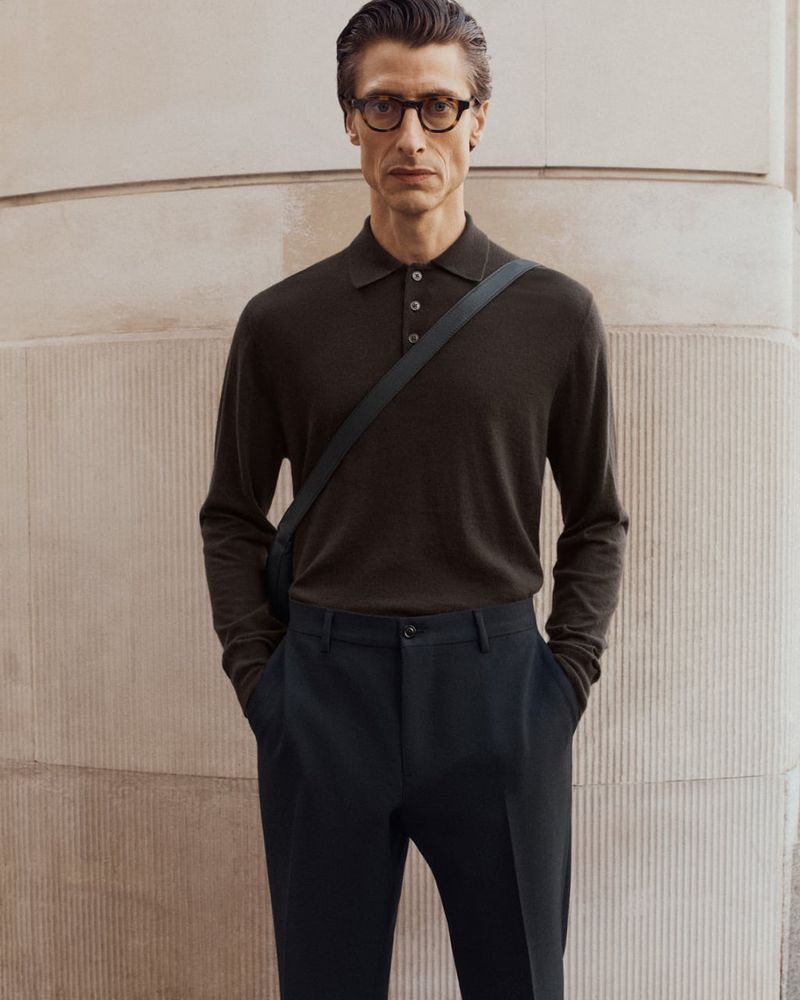A stylish adult man wearing glasses, a polo shirt, and dark pants poses against a wall, showcasing a fashion-forward look.