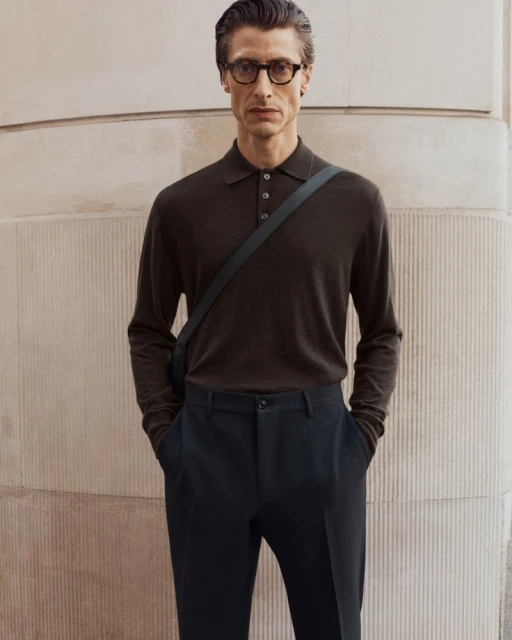 A stylish adult man wearing glasses, a polo shirt, and dark pants poses against a wall, showcasing a fashion-forward look.