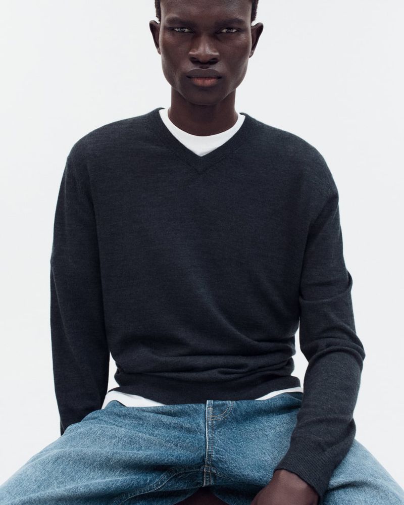 Portrait of a dark-skinned male fashion model wearing a dark grey V-neck sweater, layered over a white T-shirt and blue denim jeans.
