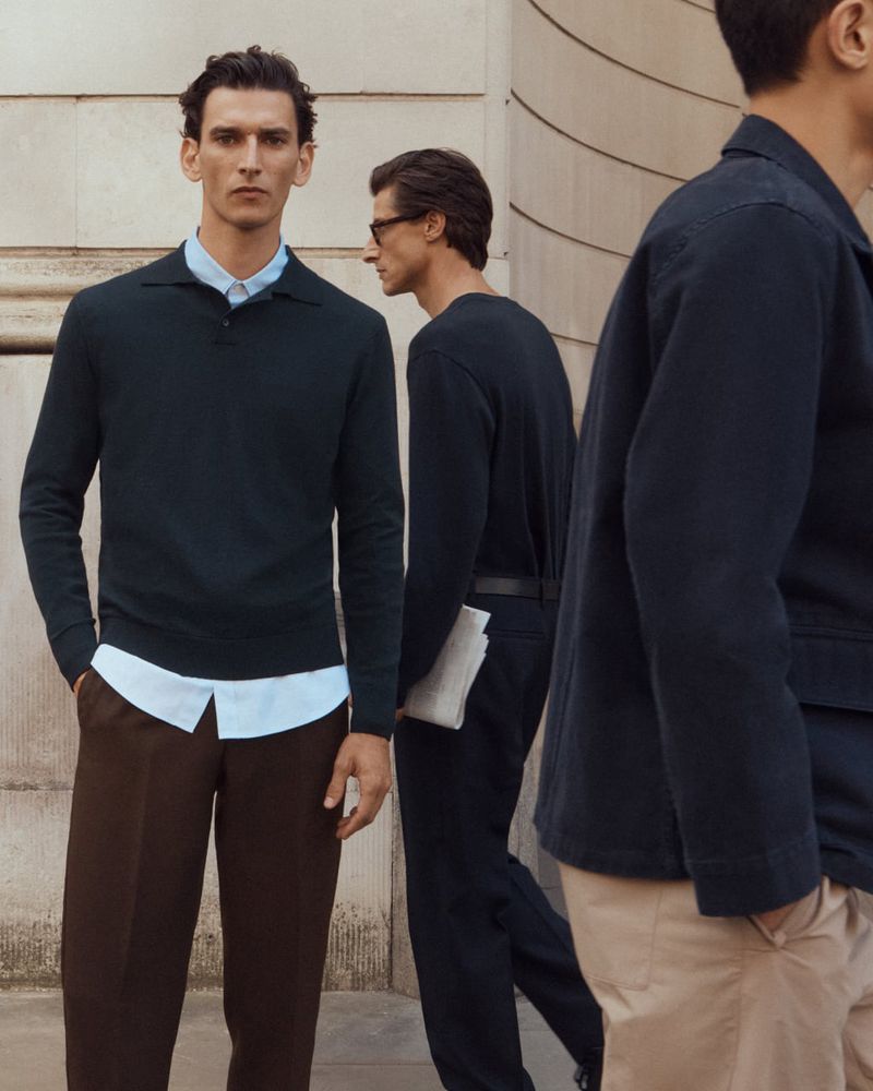 Three adult male fashion models in a neutral color palette posing outdoors on a city street in casual professional clothing.