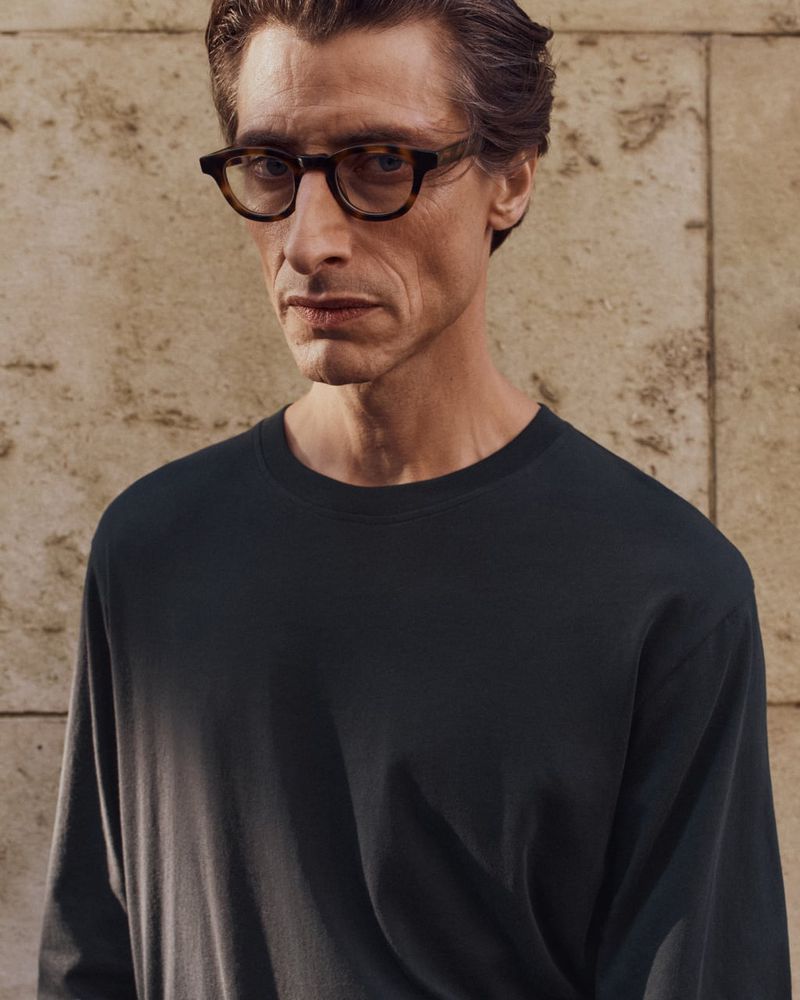 Portrait of a mature man wearing glasses and a long-sleeved black shirt, posing indoors against a textured light-brown wall.