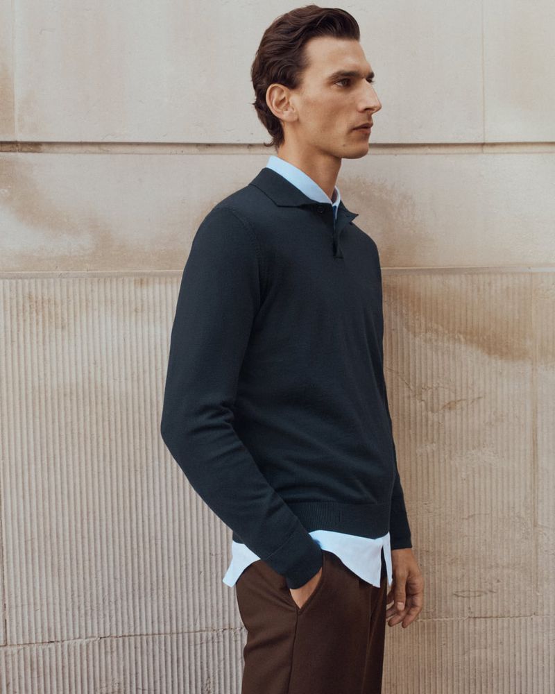A man wearing a dark polo sweater over a light blue button-down shirt against a textured stone wall.