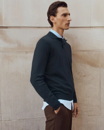 A man wearing a dark polo sweater over a light blue button-down shirt against a textured stone wall.