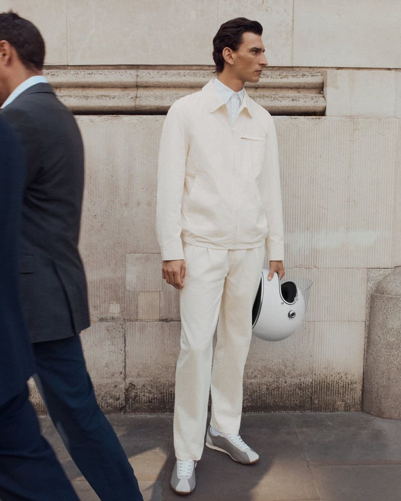 Adult man in light jacket and trousers holding white helmet stands next to passerby on urban street with neutral-toned architecture.