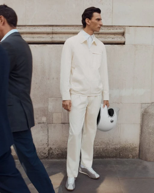 Adult man in light jacket and trousers holding white helmet stands next to passerby on urban street with neutral-toned architecture.