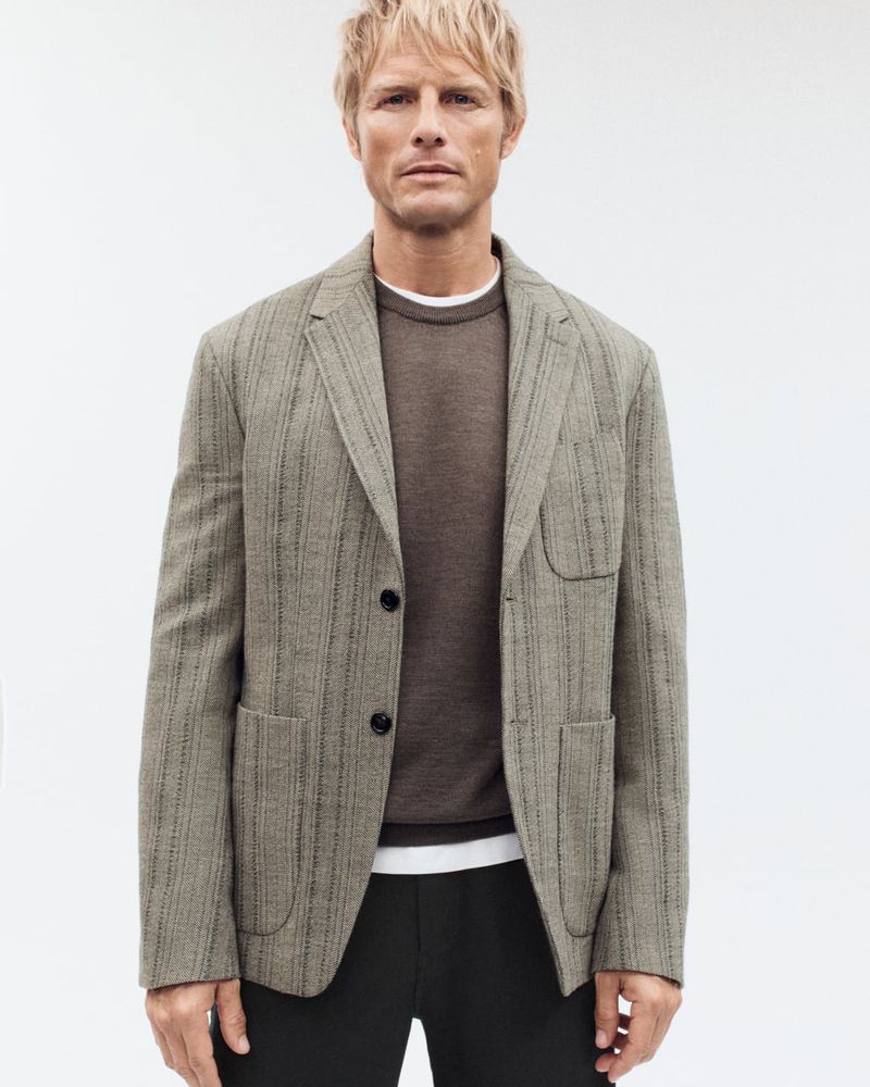 Man in tailored blazer and brown sweater poses against white backdrop in a studio shot for a fashion advertisement.