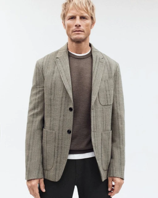 Man in tailored blazer and brown sweater poses against white backdrop in a studio shot for a fashion advertisement.
