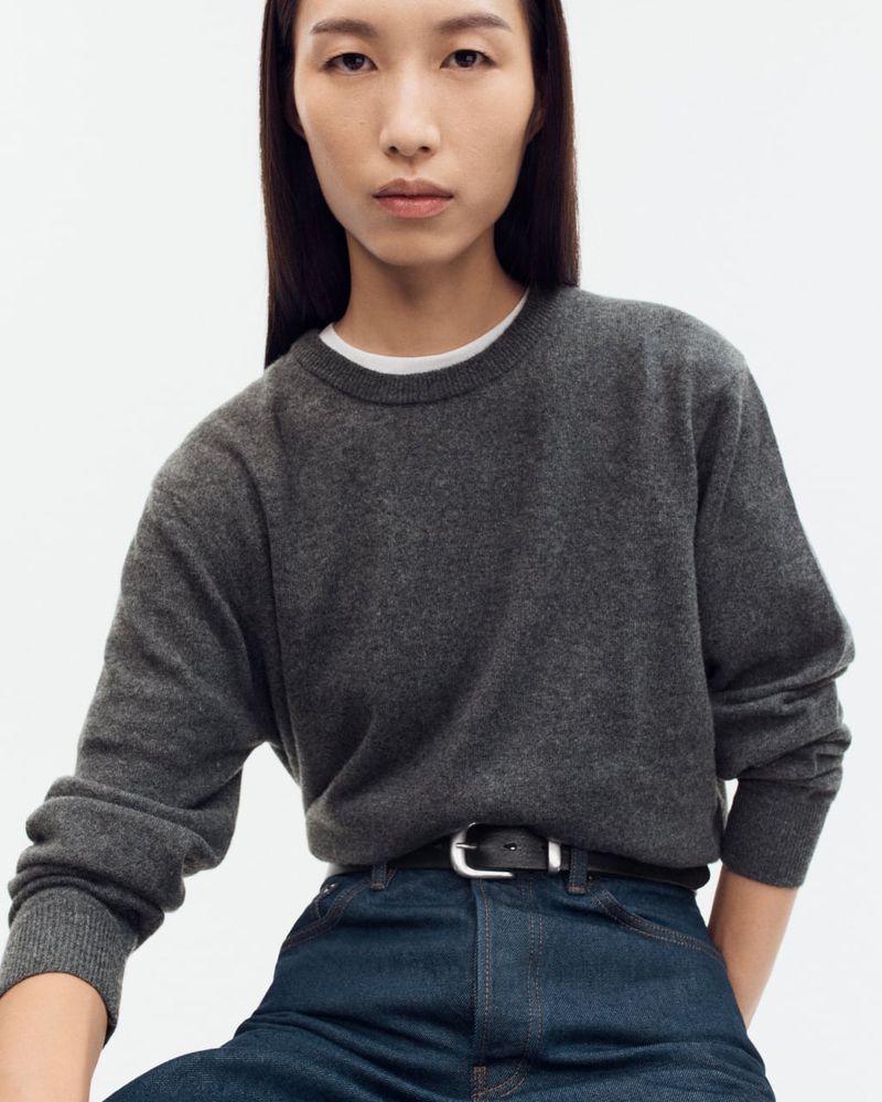 Portrait of an adult woman wearing a grey sweater and jeans with a black belt, against a white backdrop in a studio setting.