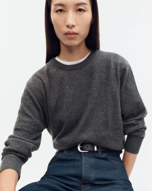 Portrait of an adult woman wearing a grey sweater and jeans with a black belt, against a white backdrop in a studio setting.