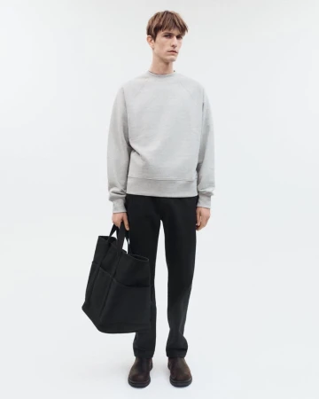 Full-length portrait of a male model wearing a grey crewneck sweatshirt and black trousers, holding a large black tote bag.