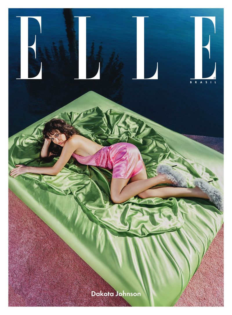 Magazine cover shot of Dakota Johnson lying on a bed covered in a shiny, bright green sheet, wearing a pink dress and furry slippers.