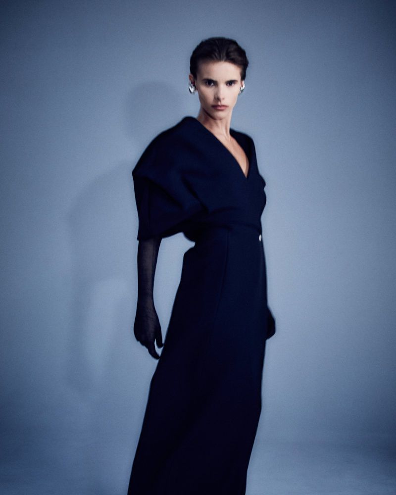 A poised fashion model in a full-length navy dress with voluminous sleeves and sheer black gloves stands against a light blue backdrop.