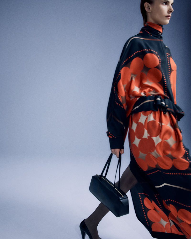 Fashionable adult female strides in a mod-print long dress with black heels and a structured handbag against a muted background.