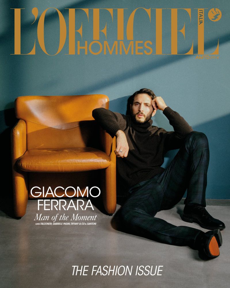 Fashion magazine cover featuring a man with a beard, wearing a dark turtleneck and jeans, sitting on the floor next to an orange chair.