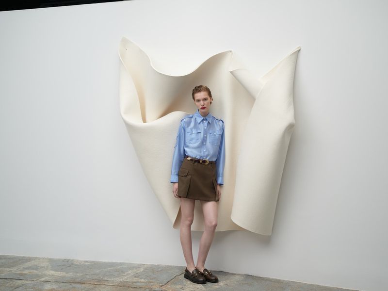 Fashion shot of a female model in a blue shirt and brown skirt, standing in front of a white backdrop in a minimalist studio setting.