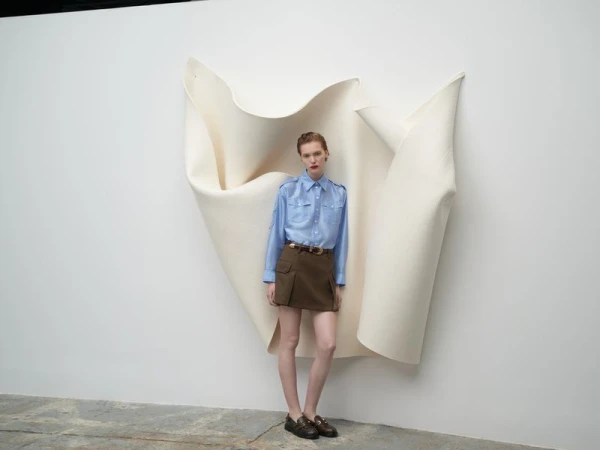 Fashion shot of a female model in a blue shirt and brown skirt, standing in front of a white backdrop in a minimalist studio setting.