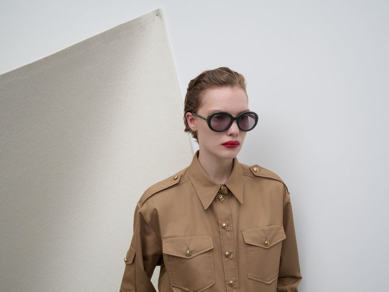 Woman in tan utility-style shirt and sunglasses against a white wall and corner backdrop in a fashion or beauty context. 