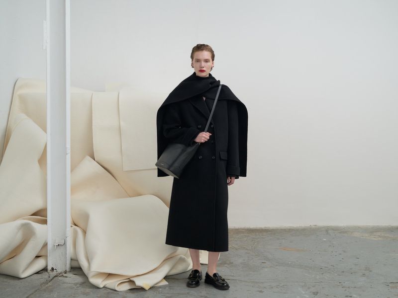 Full-length studio shot: woman in a black coat, black bag on a strap, black shoes, red lipstick, against a neutral wall and white cloth backdrop.