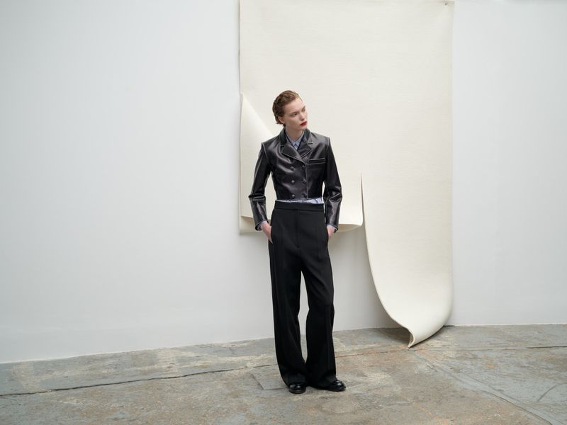 Woman in a short black leather jacket and wide black pants poses against a draped cream background in a studio setting.
