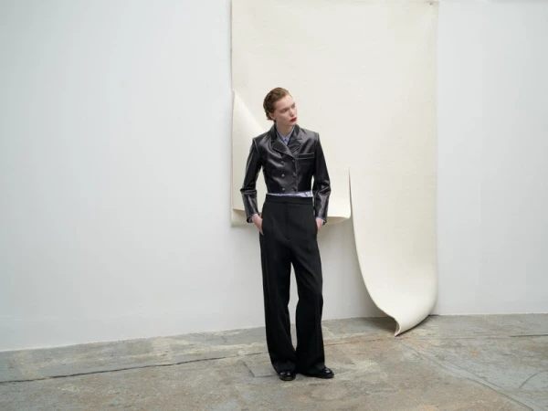 Woman in a short black leather jacket and wide black pants poses against a draped cream background in a studio setting.