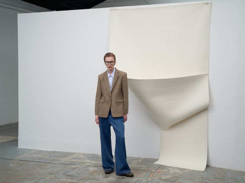 An adult woman stands in a photo studio with a tan blazer, blue shirt, baggy jeans, and brown shoes, posing in front of a white backdrop.