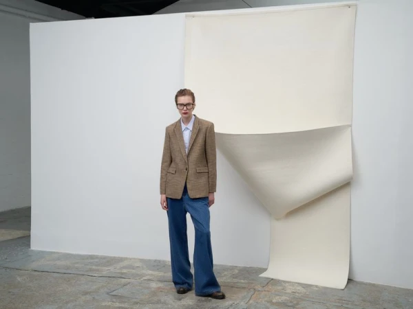 An adult woman stands in a photo studio with a tan blazer, blue shirt, baggy jeans, and brown shoes, posing in front of a white backdrop.