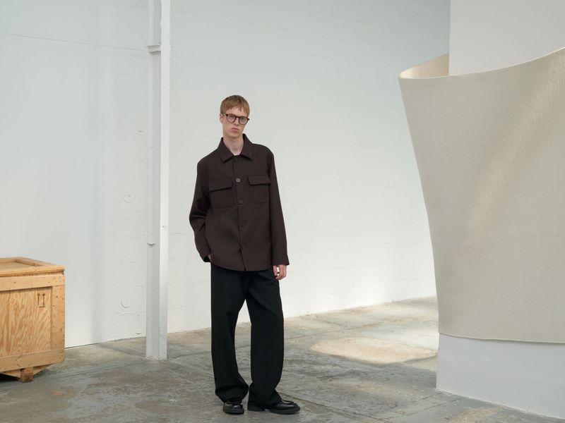 A tall, slender adult male model wearing glasses and a dark brown shirt jacket stands in a bright studio with an art installation.