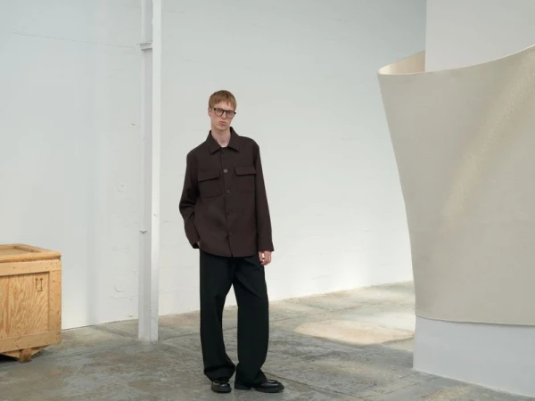 A tall, slender adult male model wearing glasses and a dark brown shirt jacket stands in a bright studio with an art installation.
