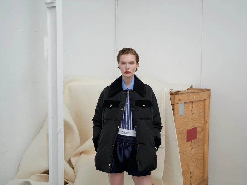A young woman poses in a black puffer jacket with dark blue shorts in a minimalist indoor setting with a wooden crate and white walls.