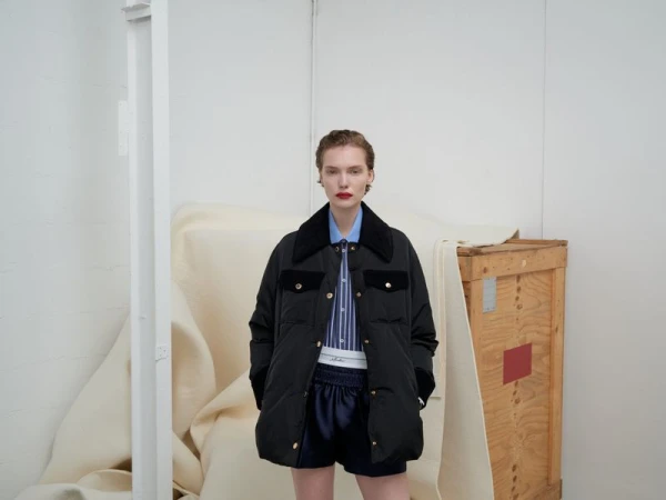 A young woman poses in a black puffer jacket with dark blue shorts in a minimalist indoor setting with a wooden crate and white walls.