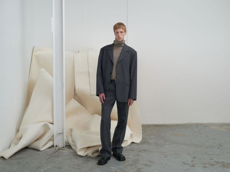 Man in grey suit with neutral turtleneck stands in a studio. Suit is slightly oversized, black leather shoes, neutral studio background.