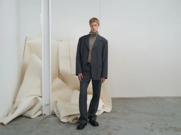 Man in grey suit with neutral turtleneck stands in a studio. Suit is slightly oversized, black leather shoes, neutral studio background.