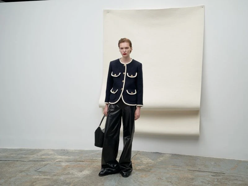 A stylish adult female with short red hair in a navy jacket with leather pants stands in a studio with a white backdrop and soft lighting.